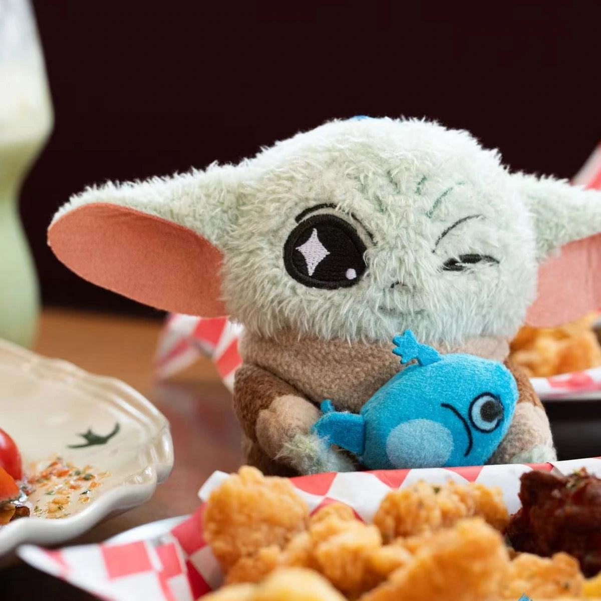 Winking Grogu plush holding a blue frog creature beside fried food