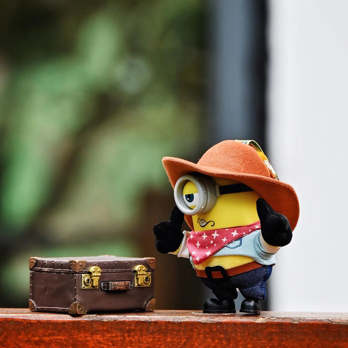 Cowboy James Minion plush standing beside a small brown suitcase