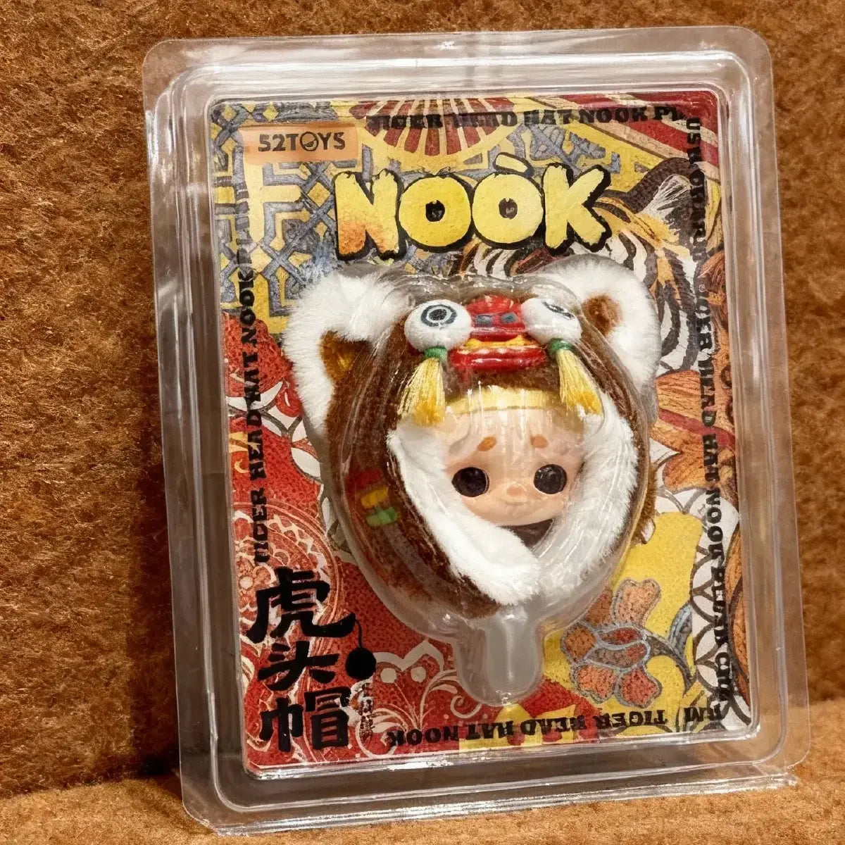 NOOK tiger-head hat plush charm clamshell package viewed at an angle on brown fabric