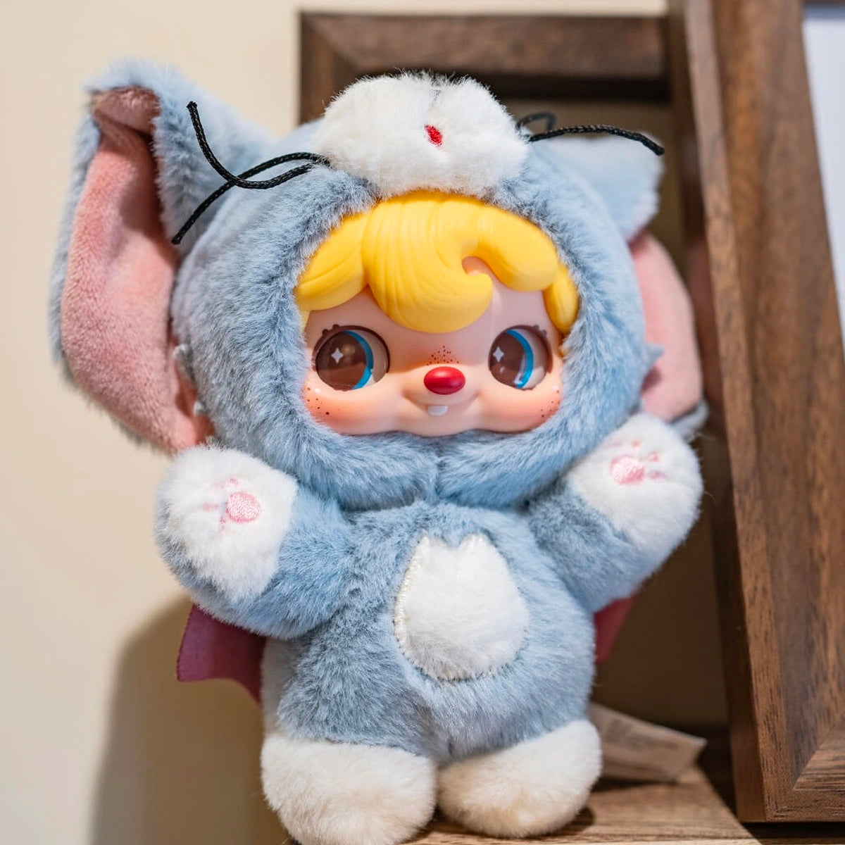 POUKAPOUKA in a gray Tom costume raising both paw gloves beside a wooden frame