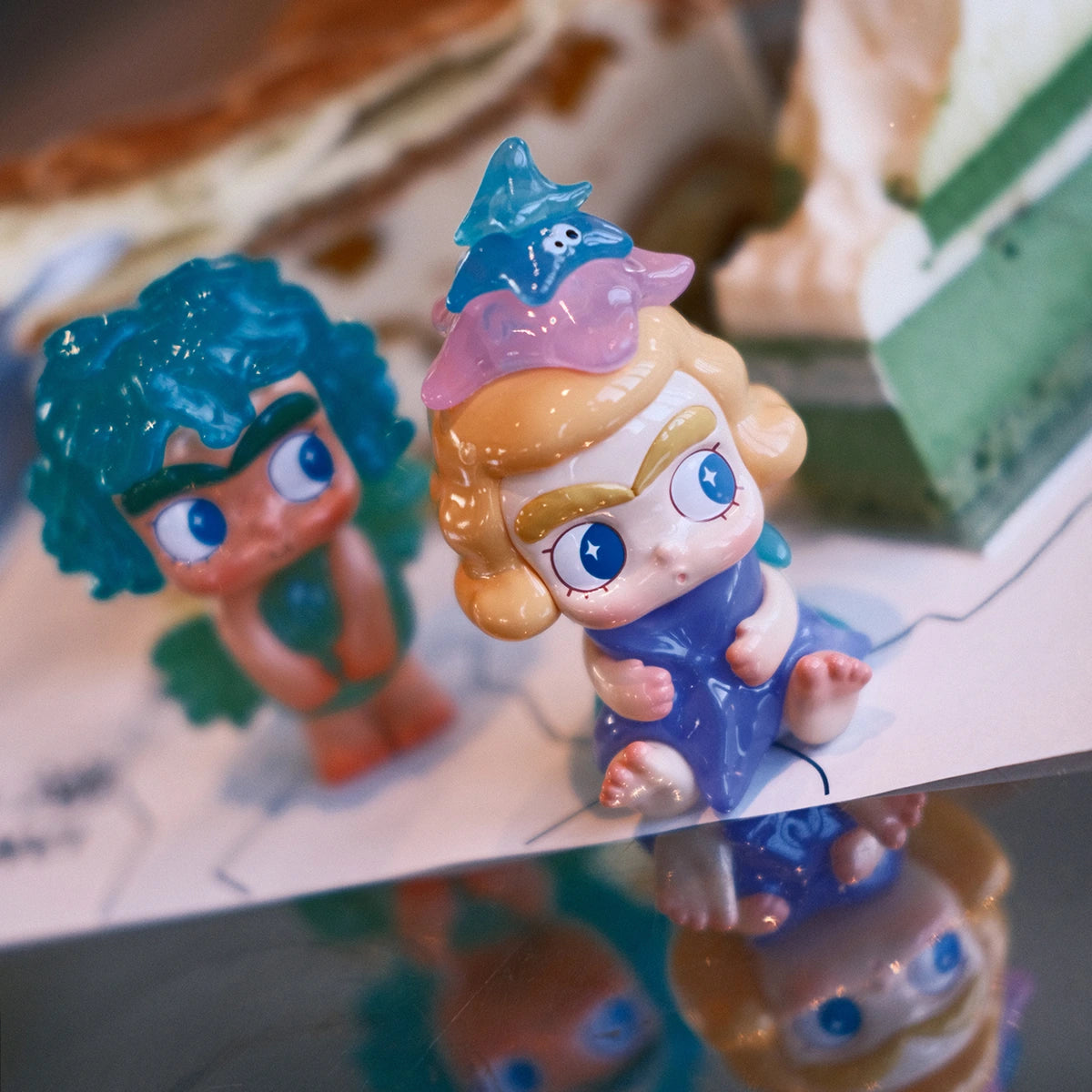 Green-haired seaweed CiCiLu and blonde starfish-costume CiCiLu posed on a reflective surface