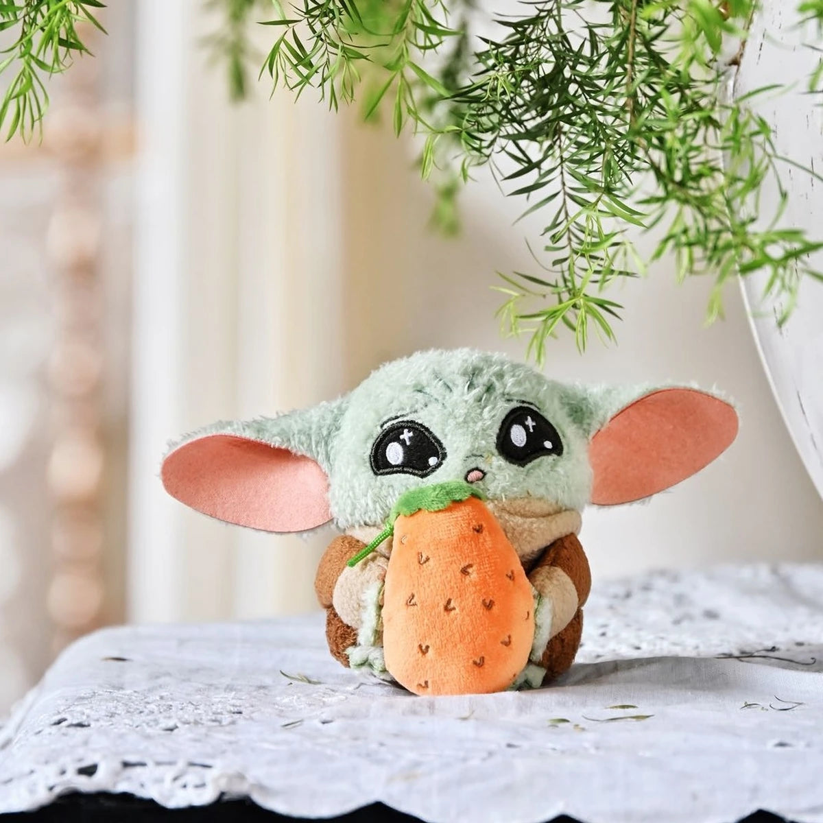 Grogu plush holding an oversized orange carrot on a lace-covered table