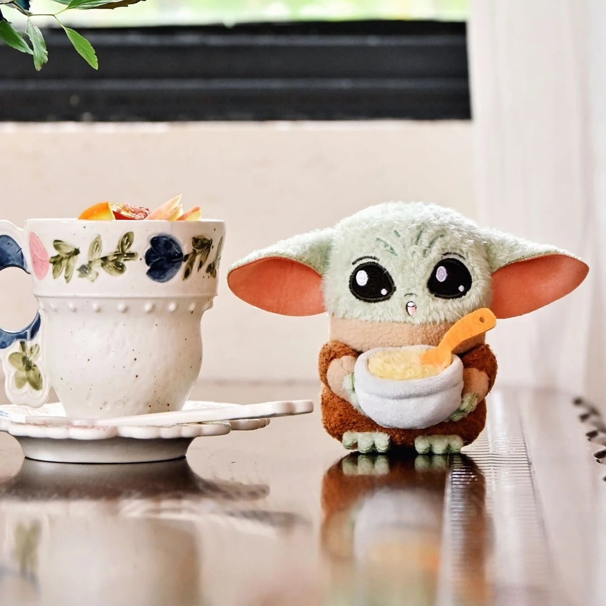 Grogu plush holding a bowl and spoon beside a floral teacup