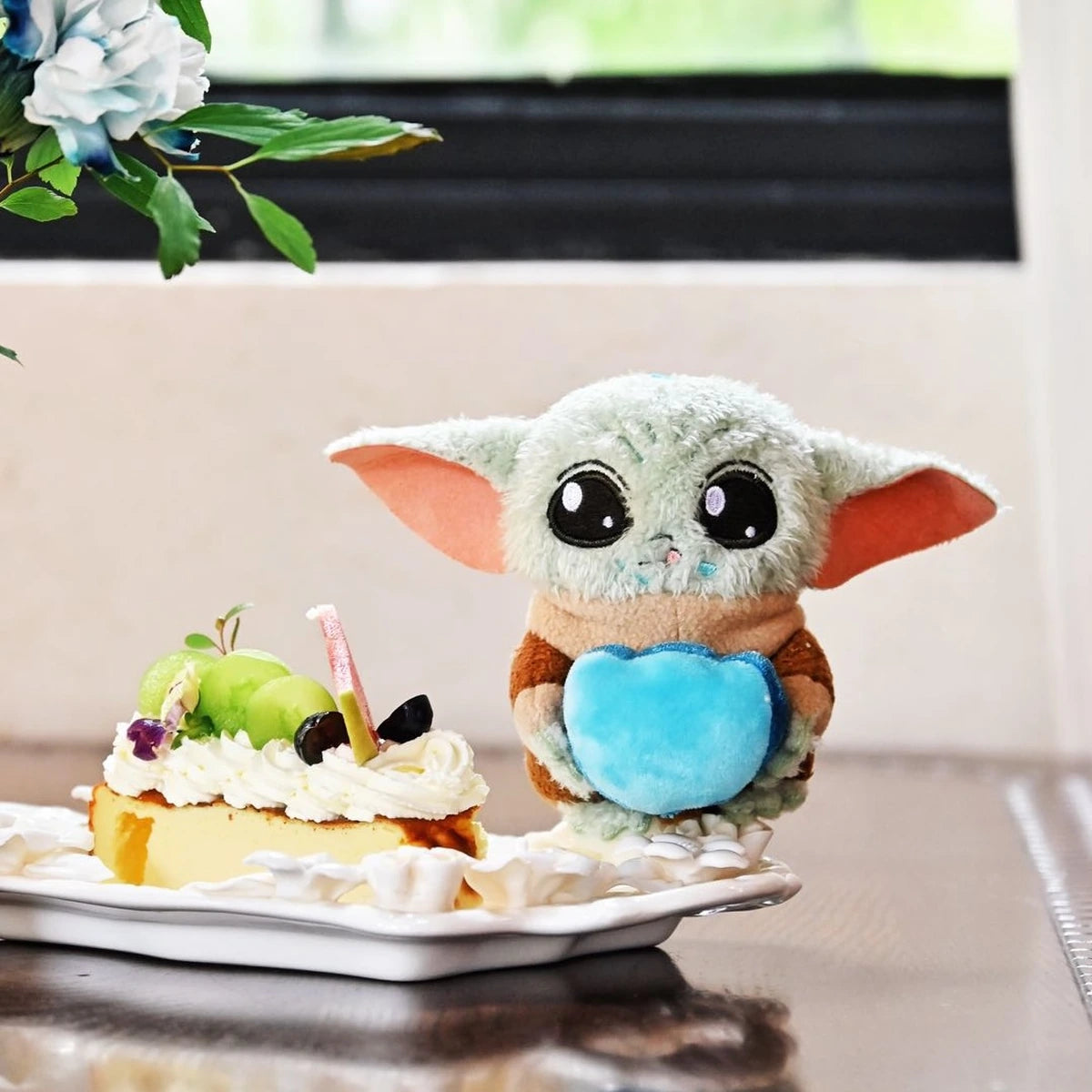 Grogu plush holding a blue heart beside a slice of fruit-topped cake