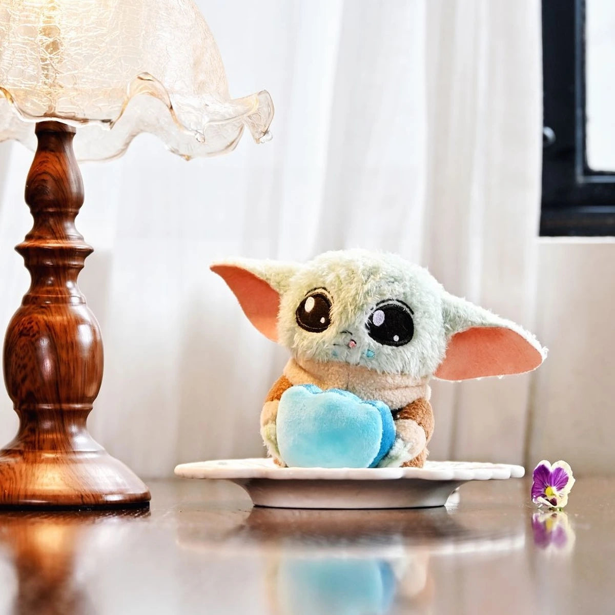 Grogu blue-heart plush seated on a white plate beside a vintage table lamp