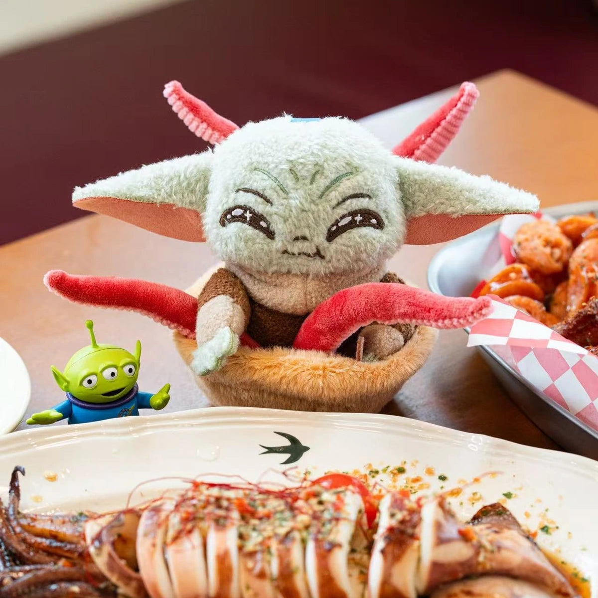 Smiling Grogu tentacle-basket plush beside a seafood meal and Toy Story Alien figure