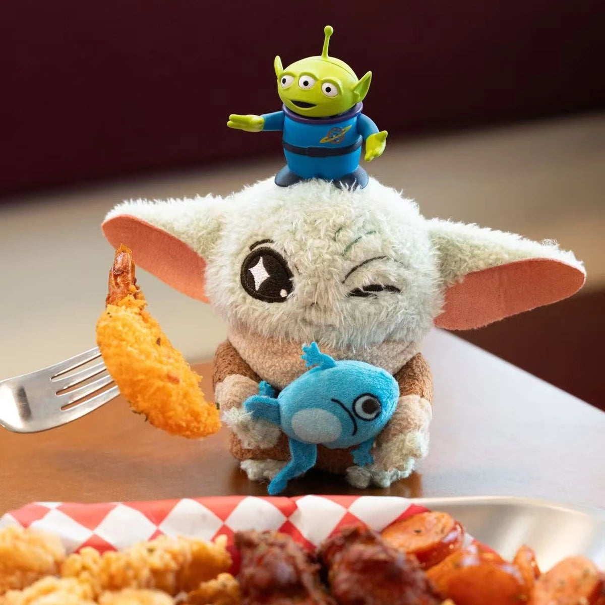 Winking Grogu with blue frog plush and Toy Story Alien figure beside a plate of fried food