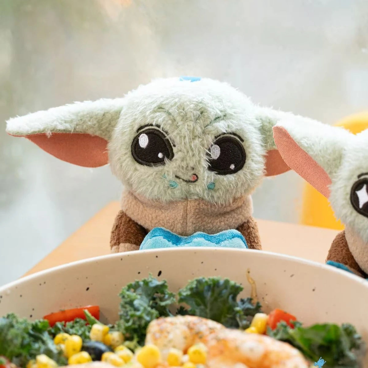 Grogu blue-heart plush peeking over a bowl of salad at a restaurant