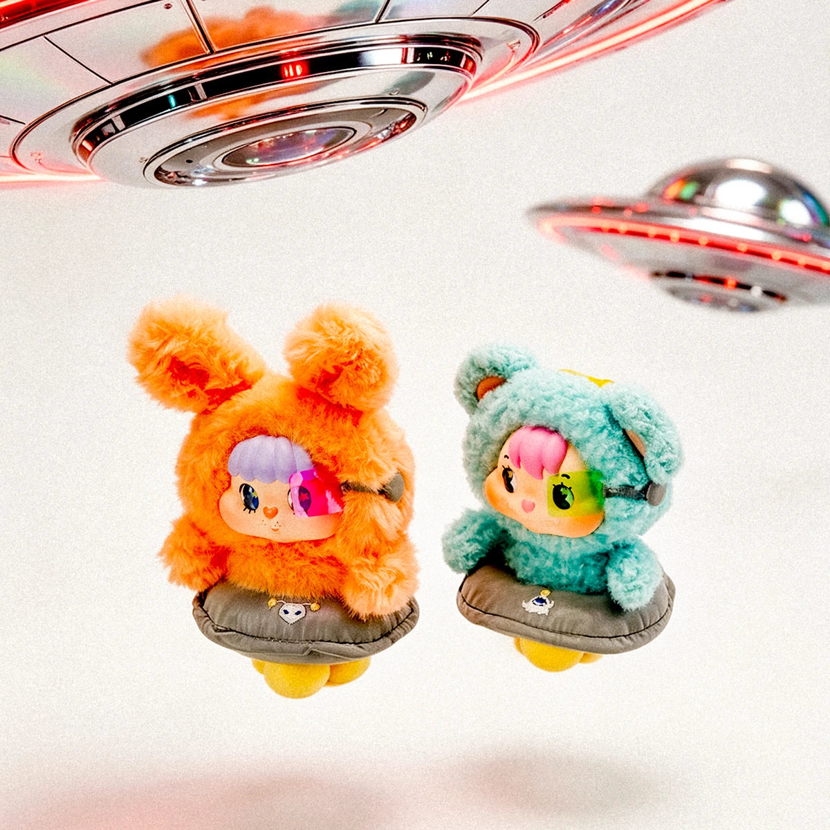 Orange rabbit and teal bear Little Buns flying-saucer plush characters beneath UFO props