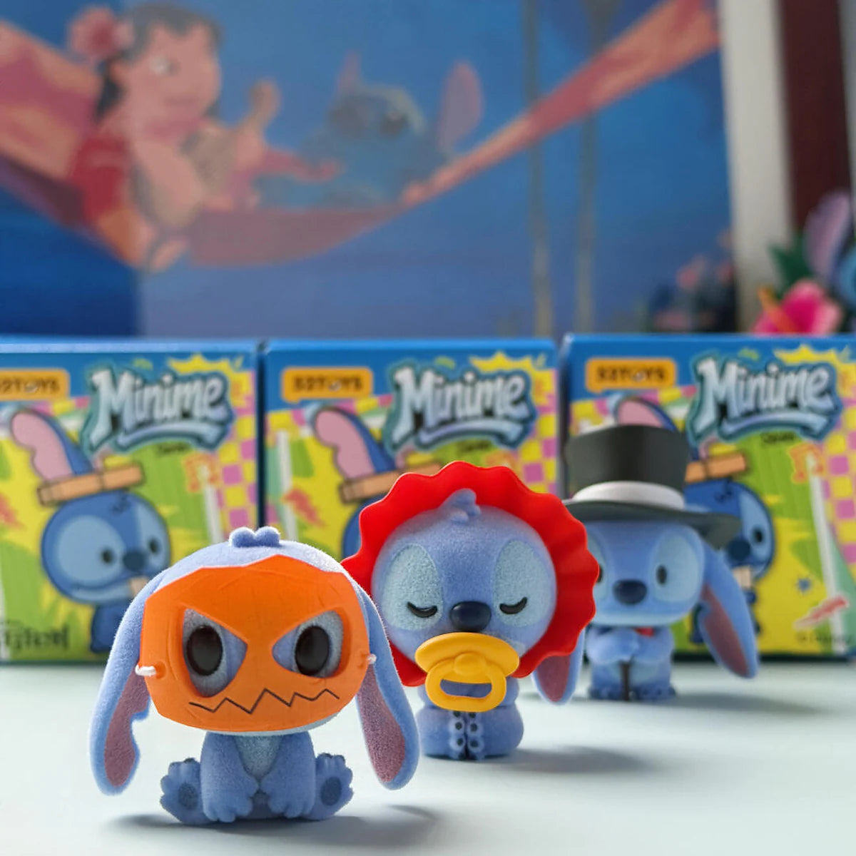 ブラインドボックスの前に並ぶ、オレンジ色のマスク、おしゃぶり付きフード、シルクハット姿のStitch MiniMeフィギュア
