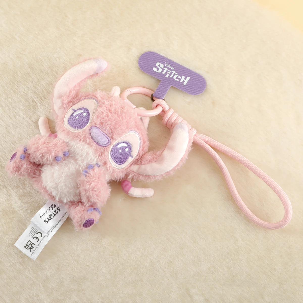 Wide-eyed pink Angel plush phone charm lying on a cream fabric background